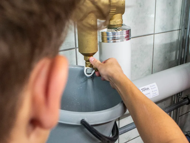 Monteur an einer Messingarmatur über einem Eimer, am Rohr ein Schick-Aufkleber